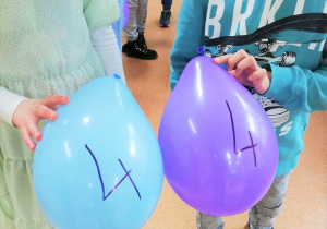 W sali lekcyjnej grupa uczniów trzyma w dłoniach kolorowe balony. Na każdym z balonów napisana jest liczba.